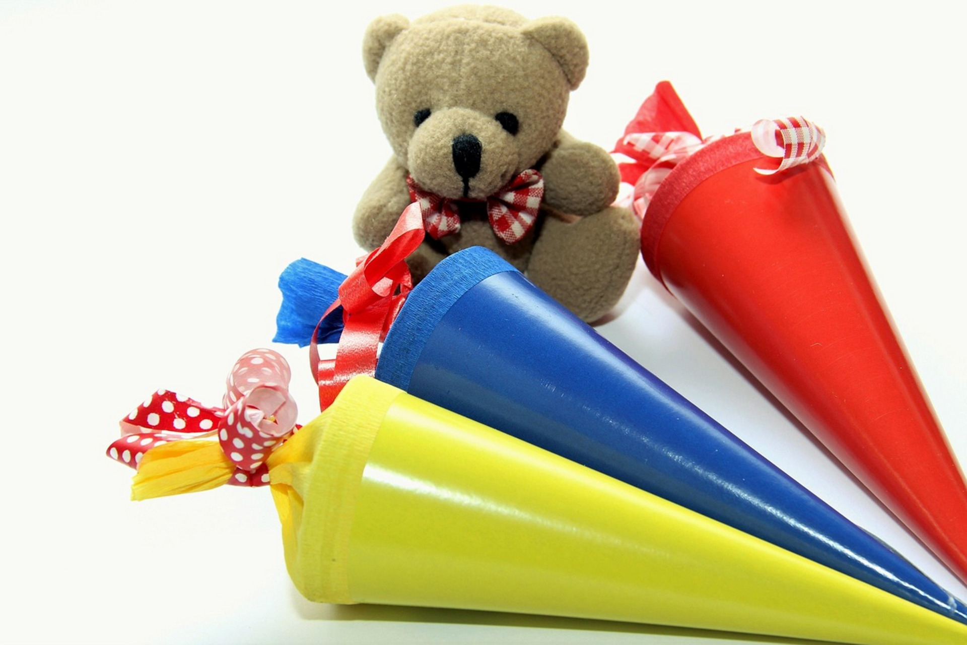 Ein kleines Stoffbärchen sitzt zwischen drei bunten Schultüten in Rot, Blau und Gelb. Jede Tüte ist mit bunten Schleifen verziert. Das Bild vermittelt ein Gefühl von Freude und Vorbereitung auf den Schulanfang.