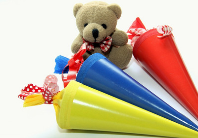 Ein kleines Stoffbärchen sitzt zwischen drei bunten Schultüten in Rot, Blau und Gelb. Jede Tüte ist mit bunten Schleifen verziert. Das Bild vermittelt ein Gefühl von Freude und Vorbereitung auf den Schulanfang.