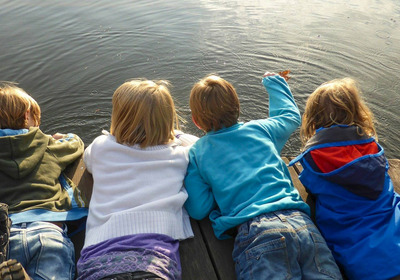 Vier Kinder liegen auf einem Holzsteg und blicken auf die Wasseroberfläche. Die Kinder tragen bunte Kleidung und haben unterschiedliche Haarlängen sowie Farben.