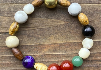 Ein Armband aus unterschiedlich gefärbten Perlen, darunter goldene, schwarze, weiße, lila, rote und grüne Steine, liegt auf einer Holzoberfläche. Die Perlen variieren in Form und Größe und sind abwechselnd angeordnet.