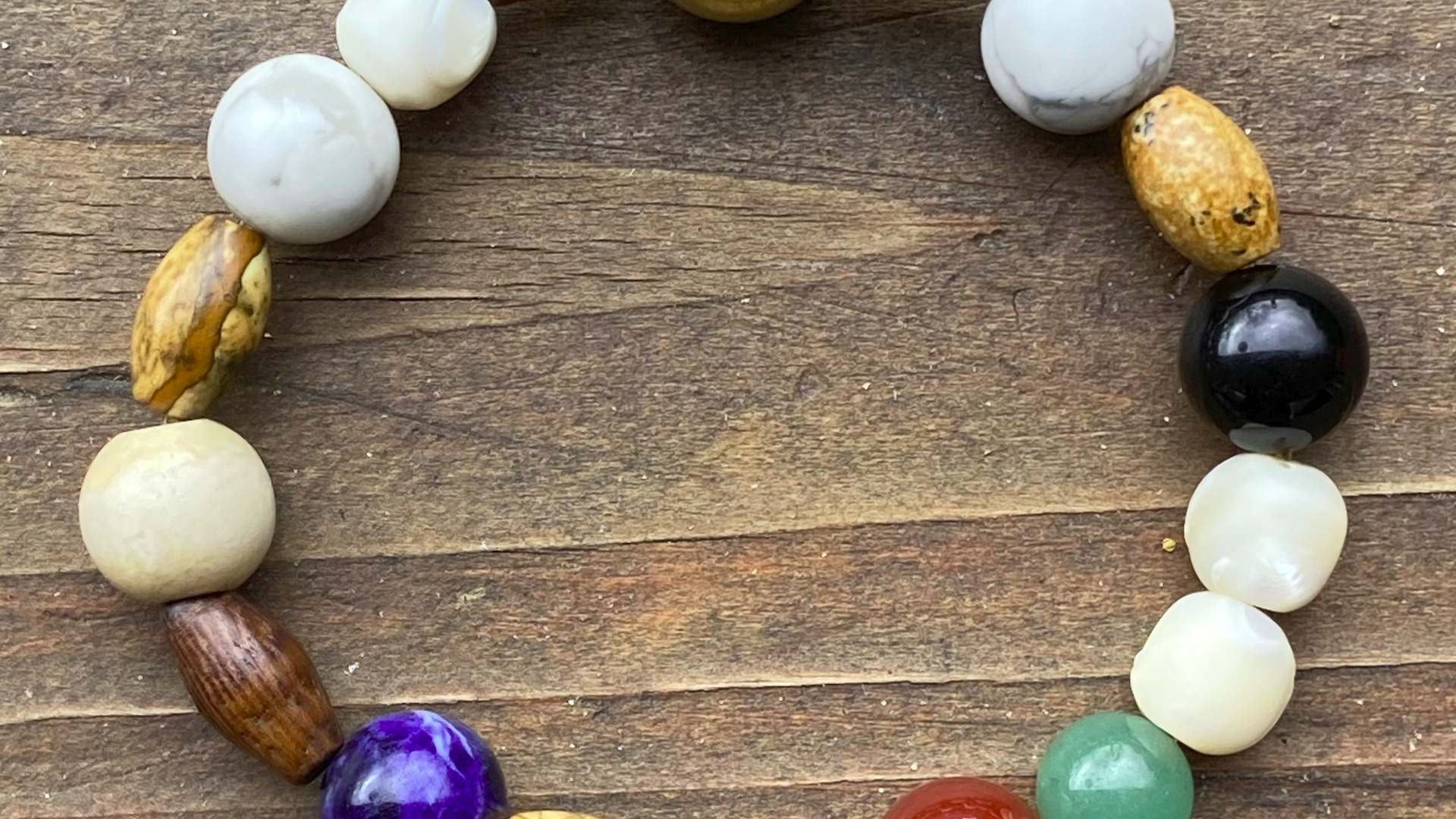 Ein Armband aus unterschiedlich gefärbten Perlen, darunter goldene, schwarze, weiße, lila, rote und grüne Steine, liegt auf einer Holzoberfläche. Die Perlen variieren in Form und Größe und sind abwechselnd angeordnet.