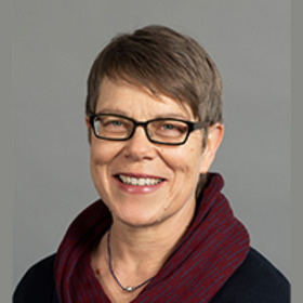Porträt einer lächelnden Frau mit kurzem, braunen Haar und einer Brille. Sie trägt einen dunklen Pullover mit einem roten Schal. Der Hintergrund ist neutral grau.