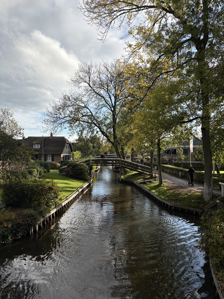 Giethoorn Ein ruhiger Wasserkanal fließt durch eine grüne Landschaft mit Bäumen und gepflegten Gärten. Eine kleine Holzbrücke überspannt den Kanal, und Personen spazieren entlang der Uferwege. Der Himmel ist bewölkt, was eine friedliche Atmosphäre schafft.