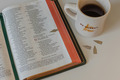 Eine geöffnete Bibel mit markierten Seiten liegt auf einem Tisch. Daneben steht eine weiße Tasse mit dem Aufdruck "BE HONEST." Ein paar kleine Papierschnipsel liegen ebenfalls auf dem Tisch, und die Tasse enthält schwarzen Kaffee.