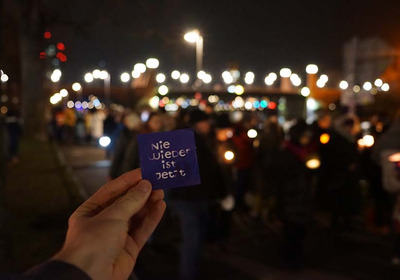 Das Foto zeigt eine Lichterdemo unscharf im Hintergrund. Im Vordergrund hält jemand ein Schild hoch mit der Aufschrift "Nie wieder ist jetzt"