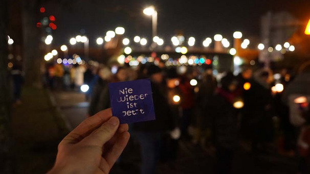 Das Foto zeigt eine Lichterdemo unscharf im Hintergrund. Im Vordergrund hält jemand ein Schild hoch mit der Aufschrift "Nie wieder ist jetzt"