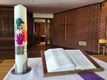 Eine Kerze mit bunten Handabdrücken und der Jahreszahl 2023 steht auf einem Tisch neben einer geöffneten Bibel. Im Hintergrund ist eine Ziegelwand mit einem Holzkreuz sichtbar. Der Raum vermittelt eine ruhige, religiöse Atmosphäre.