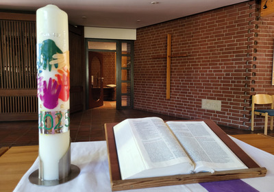 Eine Kerze mit bunten Handabdrücken und der Jahreszahl 2023 steht auf einem Tisch neben einer geöffneten Bibel. Im Hintergrund ist eine Ziegelwand mit einem Holzkreuz sichtbar. Der Raum vermittelt eine ruhige, religiöse Atmosphäre.