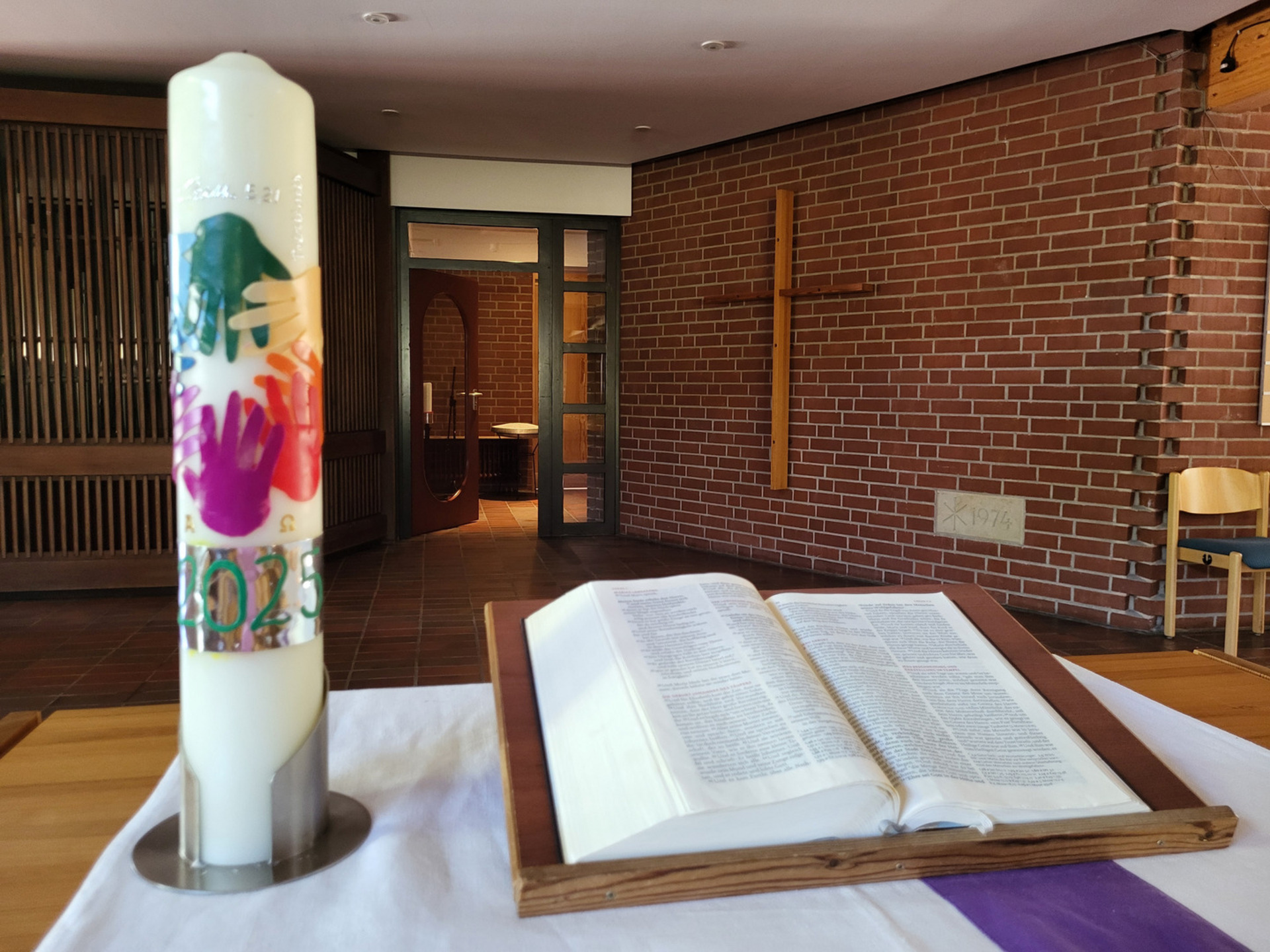 Eine Kerze mit bunten Handabdrücken und der Jahreszahl 2023 steht auf einem Tisch neben einer geöffneten Bibel. Im Hintergrund ist eine Ziegelwand mit einem Holzkreuz sichtbar. Der Raum vermittelt eine ruhige, religiöse Atmosphäre.