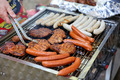 Ein Grill mit verschiedenen Fleischsorten wird zubereitet. Gegrillte Würstchen und mariniertes Fleisch sind zu sehen. Eine Hand hält eine Zange, um das Fleisch zu wenden. Der Grill ist rauchig, und der Hintergrund deutet auf eine entspannte Grillumgebung hin.