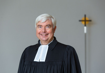 Ein lächelnder Mann mit kurzen, grauen Haaren trägt ein schwarzes Gewand mit einer weißen Halskrause. Er steht vor einer neutralen Wand, im Hintergrund ist ein Kreuz sichtbar. 