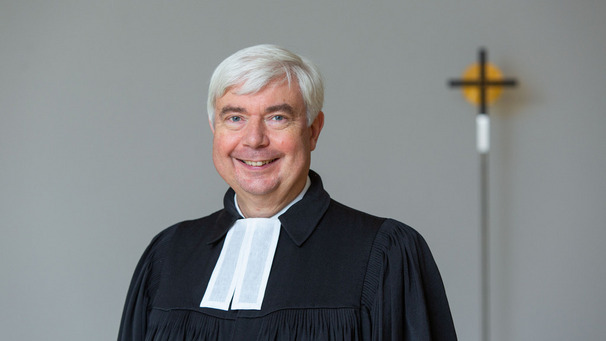 Ein Pastor in Talar steht vor einem Kreuz