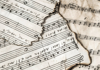 Ein Stapel abgegriffener Notenblätter liegt übereinander. Die Blätter sind vergilbt und teilweise angebrannt, mit sichtbaren Noten und Schriftzügen. Die Darstellung vermittelt einen historischen Eindruck von Musik und Notenschrift.
