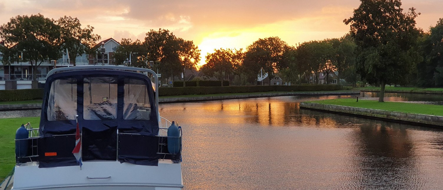 Man sieht ein Boot von hinten, welches am Ufer festgemacht ist. Im Hintergrund geht die Sonne über ein paar Häusern unter.