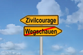 Das Bild zeigt ein Verkehrsschild mit zwei Richtungspfeilen. Der linke Pfeil weist auf "Zivilcourage" hin, während der rechte Pfeil "Wegschaunen" zeigt, aber durchgestrichen ist. Der Hintergrund besteht aus einem blauen Himmel mit Wolken.