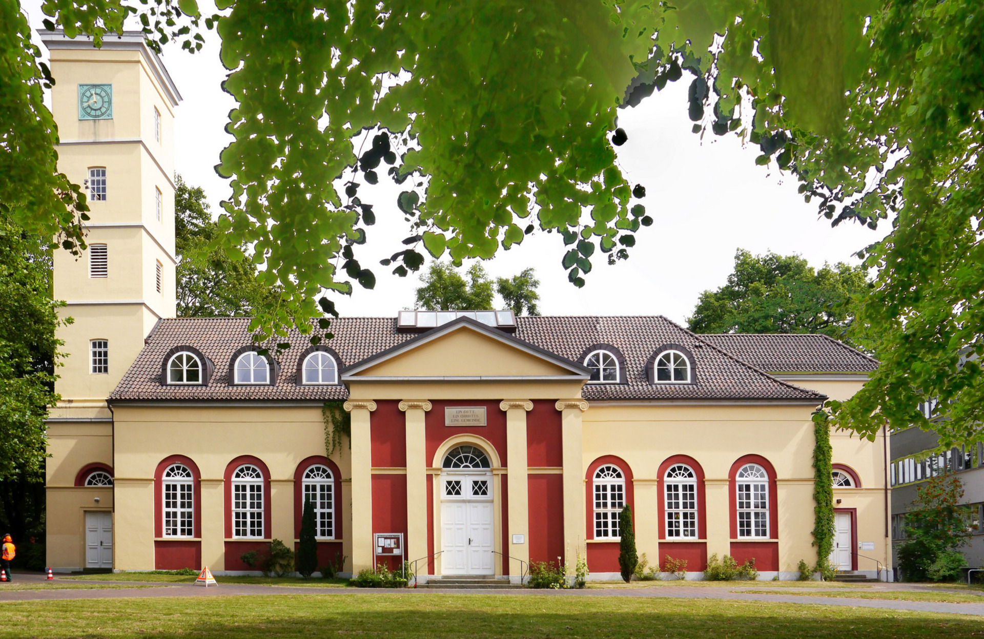 Das Bild zeigt ein historisches Gebäude mit roter und gelber Fassade, das große Fenster und einen zentralen Eingang hat. Auf der linken Seite steht ein Uhrturm, der das Gesamtbild ergänzt. Grünpflanzen umgeben das Gebäude und verleihen der Szene einen natürlichen Rahmen.
