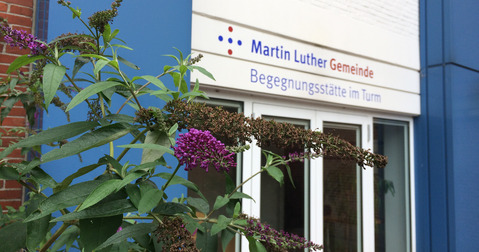 Blick auf die Terassentür zum Begegnungszentrum mit der Beschilderung über der Tür. Im Vordergrund blüht der Flieder lila.