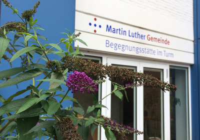 Blick auf die Terassentür zum Begegnungszentrum mit der Beschilderung über der Tür. Im Vordergrund blüht der Flieder lila.