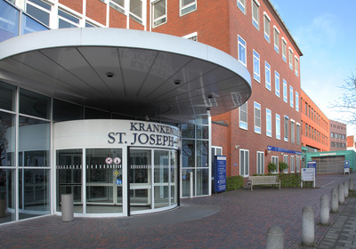 Eingang des Krankenhauses St. Joseph mit einem runden Überstand. Die Fassade ist in Ziegelrot gehalten, und große Fenster sind sichtbar. Vor dem Eingang ist ein gepflasterter Weg mit mehreren Steinpflöcken und einer Sitzbank.
