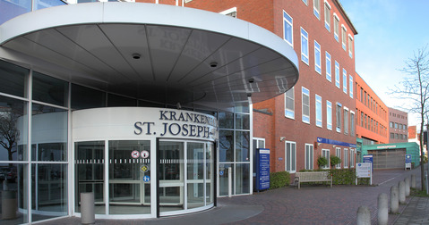 Eingang des Krankenhauses St. Joseph mit einem runden Überstand. Die Fassade ist in Ziegelrot gehalten, und große Fenster sind sichtbar. Vor dem Eingang ist ein gepflasterter Weg mit mehreren Steinpflöcken und einer Sitzbank.