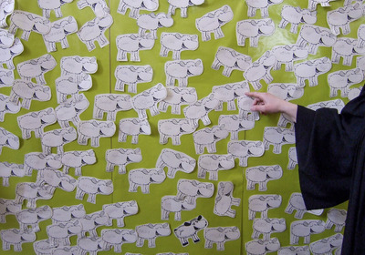 Eine bunte Wand mit vielen handgezeichneten Schafen, jedes mit einem Namen beschriftet. Eine Hand zeigt auf eines der Schafe. Der Hintergrund ist grün und die Schafe sind schwarz-weiß.