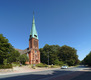 Ein rotes Backsteingebäude mit einem grünen, spitzen Kirchturm und einer Uhr an der Fassade. Umgeben von Bäumen und einem kleinen Garten. Im Vordergrund verläuft eine Straße, auf der ein weißes Auto fährt. Der Himmel ist klar und blau.