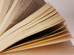 Ein offenes Buch ist im Bild zu sehen, wobei die Seiten sanft aufgefächert sind. Die Seiten sind gelblich und zeigen einen Teil des Textes, der leicht lesbar ist. Der Hintergrund ist neutral und lässt das Buch hervorstechen.
