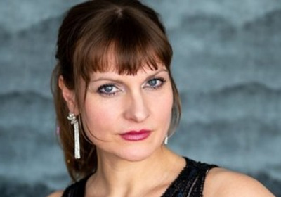 Porträt einer Frau mit mittellangen, braunen Haaren und einer eleganten Kleidung. Sie hat einen selbstbewussten Gesichtsausdruck und trägt auffälligen Schmuck. Der Hintergrund ist in sanften Grautönen gehalten.