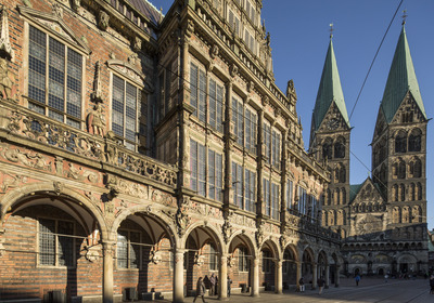 Das Bild zeigt die beeindruckende Fassade des Bremer Rathauses mit kunstvoll verzierten Balkonen und Bögen im Vordergrund. Im Hintergrund ragen die markanten Türme des Bremer St. Petri Doms in den blauen Himmel. 