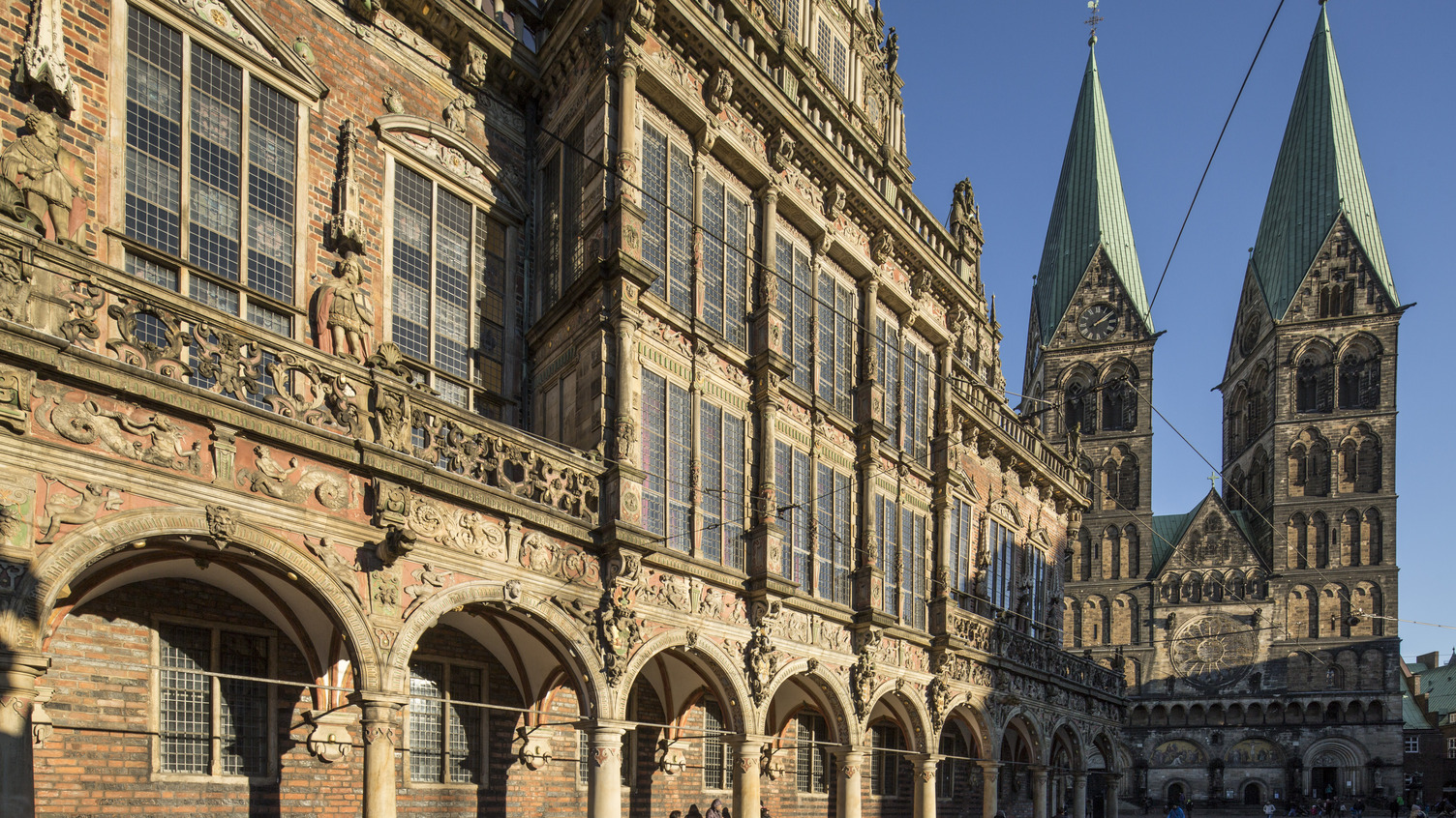 Das Bild zeigt die beeindruckende Fassade des Bremer Rathauses mit kunstvoll verzierten Balkonen und Bögen im Vordergrund. Im Hintergrund ragen die markanten Türme des Bremer St. Petri Doms in den blauen Himmel.