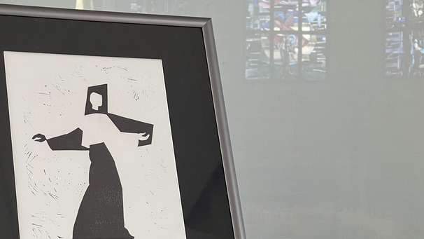 Eine stilisierte Schwarz-Weiß-Zeichnung einer Figur in einer Robe, die eine Hand ausstreckt, steht in einem Rahmen auf einem Tisch. Vor der Zeichnung liegen gefaltete Karten mit Silhouetten von Personen und informativen Texten, die sich auf das Gesamtwerk beziehen.