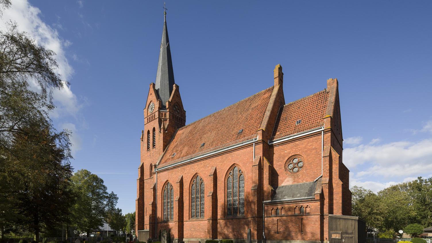 Eine rote Backsteinkirche mit einem hohen Turm und einem Ziffernblatt an der Front. Die Fassade zeigt große gotische Fenster und ein rundes Fenster. Um die Kirche herum sind ein Friedhof und Grünanlagen sichtbar, die eine ruhige Atmosphäre schaffen. Der Himmel ist blau mit einigen Wolken.