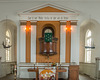 Der Innenraum einer Kirche mit einer hölzernen Kanzel und einem grünen Tuch, das Vögel zeigt. Ein Altar mit einer dekorativen Tischdecke und Blumen ist sichtbar. An den Wänden hängen kleine Schiffsmodelle, und es gibt große Fenster, die Licht hereinlassen.