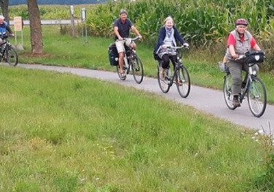 Eine Gruppe von vier Radfahrern fährt auf einem Weg entlang einer Wiese. Die Personen sind unterschiedlich gekleidet und transportieren Gepäck. Im Hintergrund sind hohe Pflanzen und ein ländliches Umfeld sichtbar.