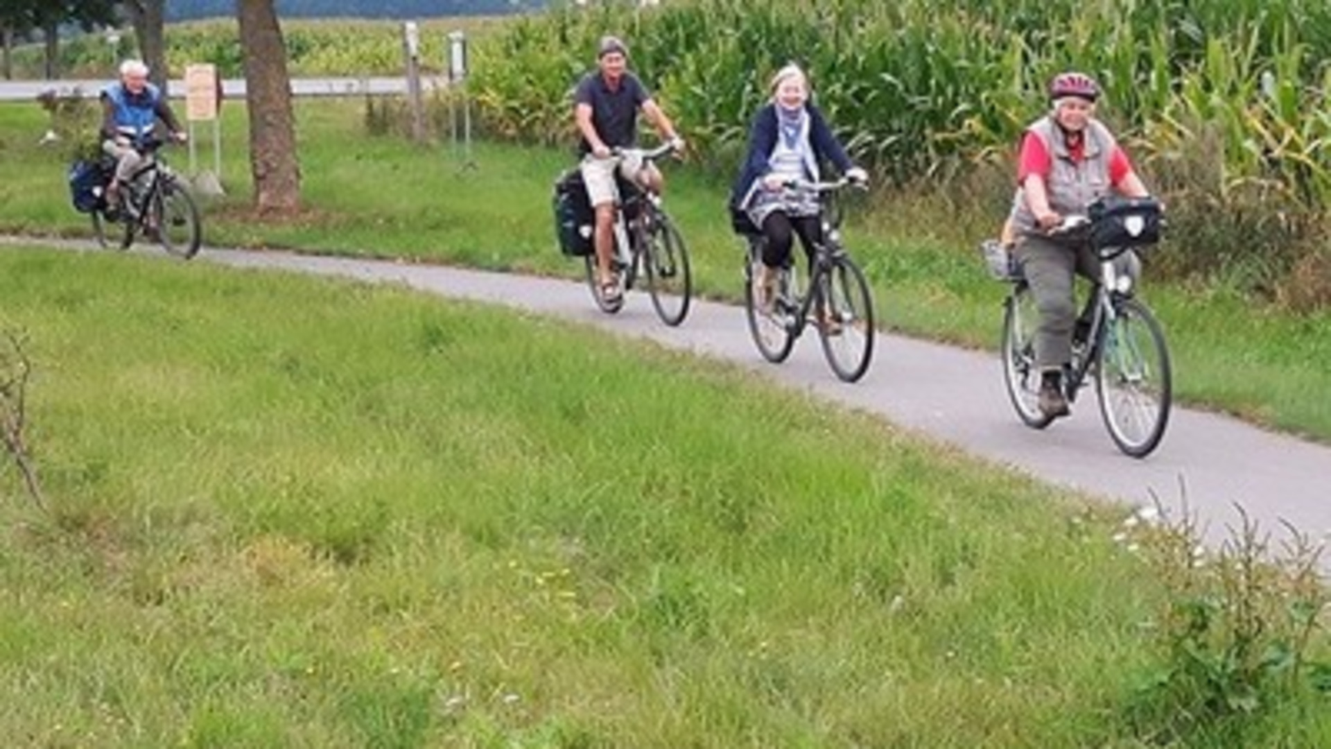 Eine Gruppe von vier Radfahrern fährt auf einem Weg entlang einer Wiese. Die Personen sind unterschiedlich gekleidet und transportieren Gepäck. Im Hintergrund sind hohe Pflanzen und ein ländliches Umfeld sichtbar.