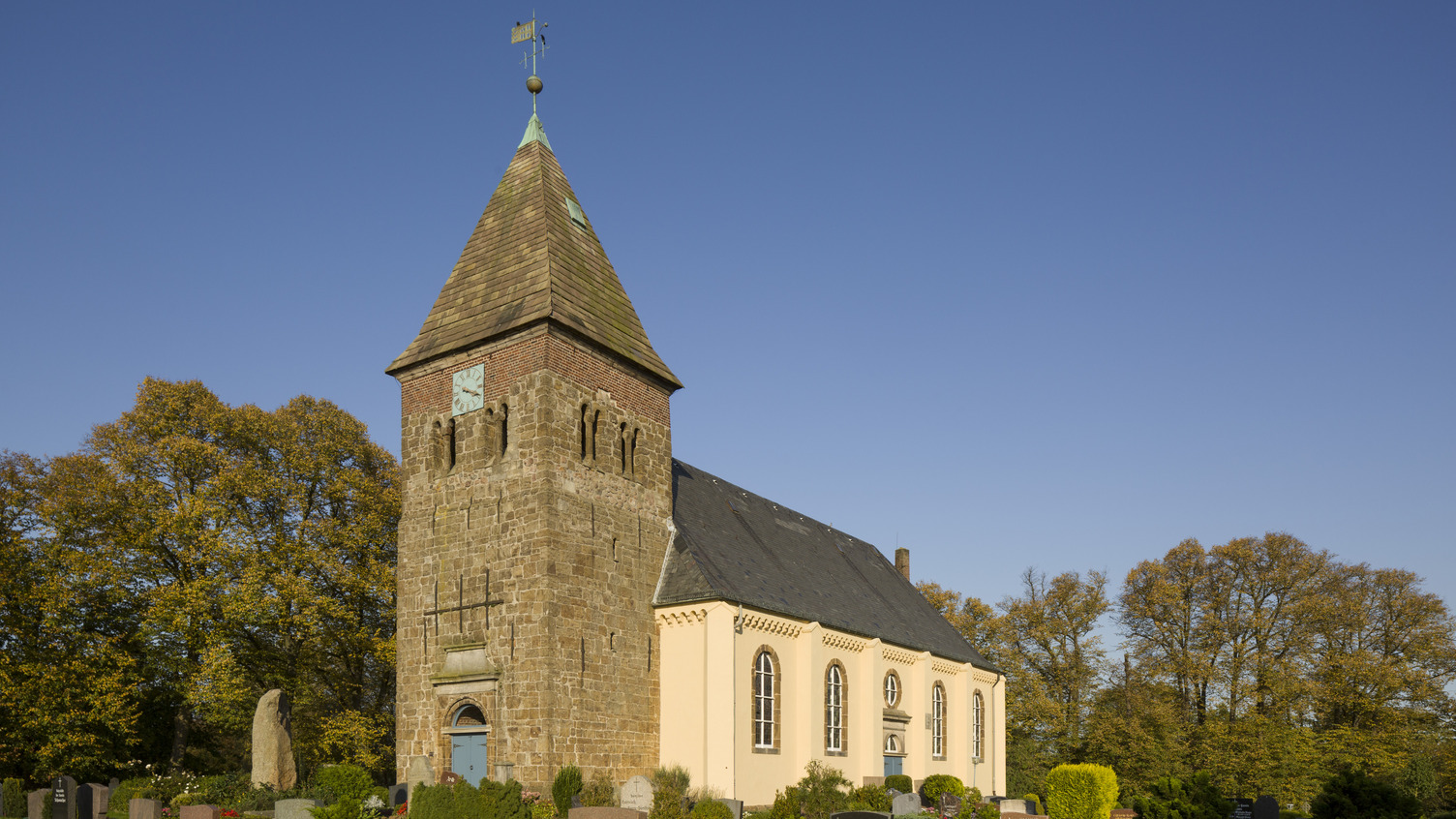 Eine alte Kirche mit einem rechteckigen Turm und einem spitzen Dach steht vor klarem blauen Himmel. Am Fuß der Kirche sind Gräber und Begrünung zu sehen. Die Architektur kombiniert Stein und Putz, mit großen Fenstern und einem Uhrenturm.