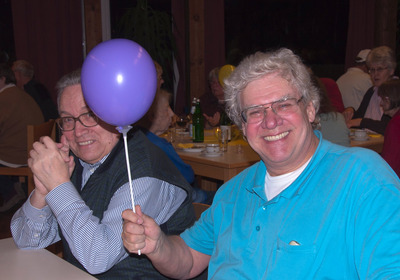 Zwei Männer sitzen lächelnd am Tisch, einer hält einen Luftballon