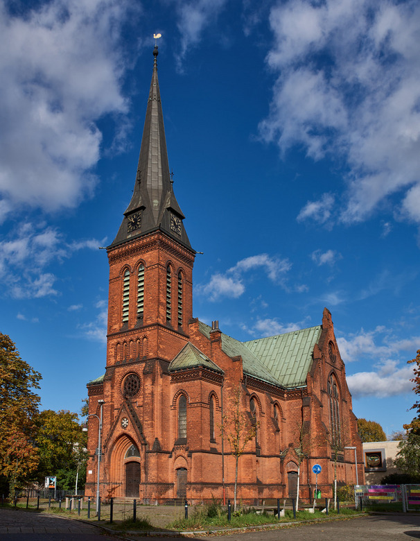 Eine rote Backsteinkirche mit einem hohen, spitzen Turm und einem grünen Satteldach. Die Fassade zeigt Fenster mit vertikalen Streben. Im Hintergrund erstreckt sich ein blauer Himmel mit einigen Wolken. Herbstlich gefärbte Bäume befinden sich im Vordergrund.