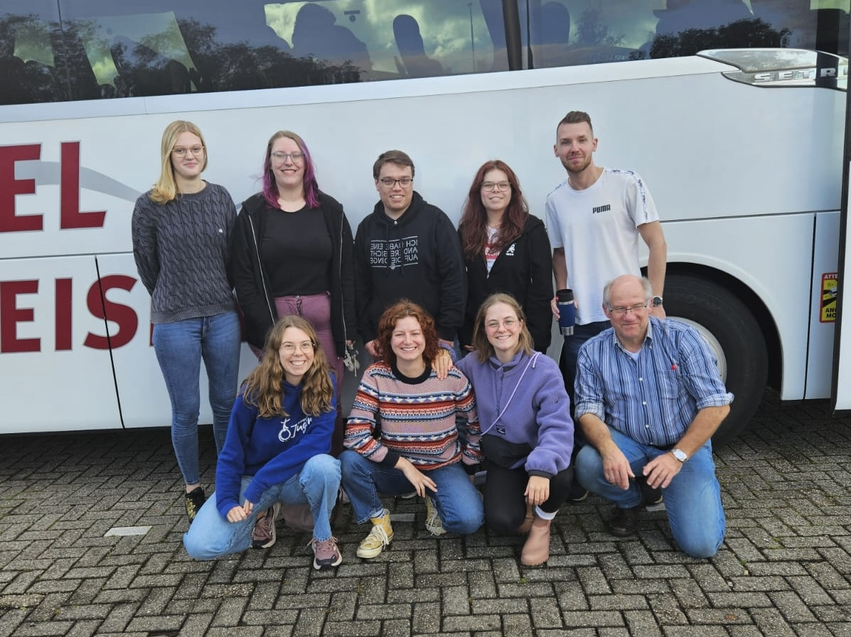 Team Eine Gruppe von neun Personen steht vor einem Reisebus. Sie posieren für das Foto, einige lächeln in die Kamera. Der Bus hat die Aufschrift „Frenzel Reisen“. Im Hintergrund ist ein wolkiger Himmel zu sehen.