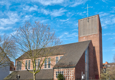 Eine moderne Kirche mit einem hohen, rechteckigen Turm und einem Kreuz an der Spitze. Die Fassade ist aus roten Ziegeln, mit großen Fenstern, die Tageslicht hereinlassen. Im Vordergrund sind kahle Bäume sichtbar, vor einem klaren blauen Himmel.