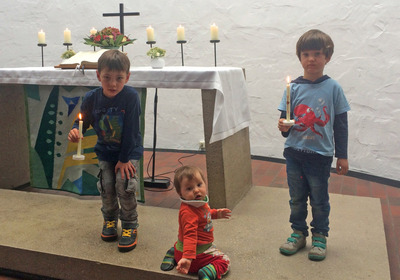 Drei Kinder stehen und sitzen mit einer Kerze vor einem Altar