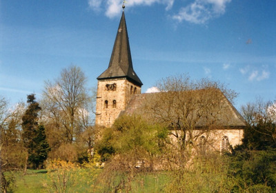 Eine historische Kirche mit einem hohen, spitzen Turm steht inmitten von Bäumen und Wiesen. Der Himmel ist blau mit wenigen Wolken. Die Architektur ist einfach, mit einem markanten Dach und kleinen Fenstern, die den Charakter der Kirche unterstreichen.