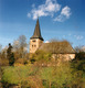 Eine historische Kirche mit einem hohen, spitzen Turm steht inmitten von Bäumen und Wiesen. Der Himmel ist blau mit wenigen Wolken. Die Architektur ist einfach, mit einem markanten Dach und kleinen Fenstern, die den Charakter der Kirche unterstreichen.