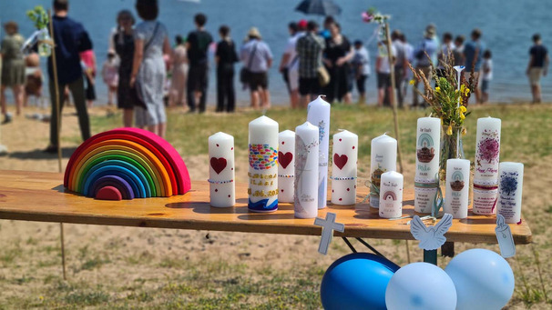 Das Bild Taufkerzen, die auf einem Tisch stehen. Im Hintergrund stehen Menschen am Fussufer