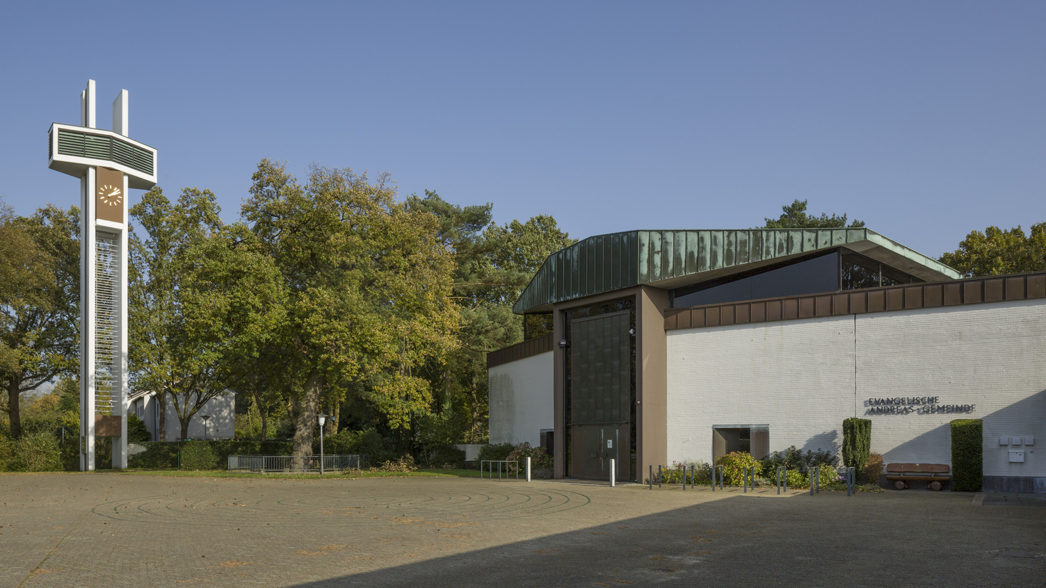 Ein modernes Gebäude mit einem auffälligen, grünen Dach und einer großen Glasfront. Vor dem Gebäude steht ein hoher, weißer Uhrturm in Form eines Kreuzes. Umgeben von Bäumen und einer gepflasterten Fläche, die leicht uneben ist. Die Fassade trägt die Aufschrift „Evangelische Andreas-Gemeinde“.