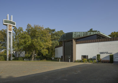 Ein modernes Gebäude mit einem auffälligen, grünen Dach und einer großen Glasfront. Vor dem Gebäude steht ein hoher, weißer Uhrturm in Form eines Kreuzes. Umgeben von Bäumen und einer gepflasterten Fläche, die leicht uneben ist. Die Fassade trägt die Aufschrift „Evangelische Andreas-Gemeinde“.
