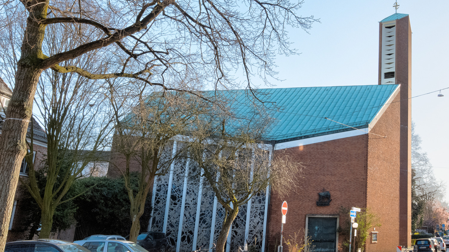 Eine moderne Kirche mit rotem Mauerwerk und einem auffälligen, grünen Blechdach. Der Turm hat eine ungewöhnliche Form mit verschiedenen Öffnungen. Umgeben von Bäumen steht das Gebäude in einer städtischen Umgebung, in der man auch parkende Autos sieht.