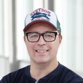 Ein Mann mit Brille und einer Cap sitzt vor einer hellen Hintergrundumgebung. Er lächelt freundlich und trägt ein dunkelblaues Oberteil. Seine Cap hat den Schriftzug "STETSON" sowie ein Tiermotiv.