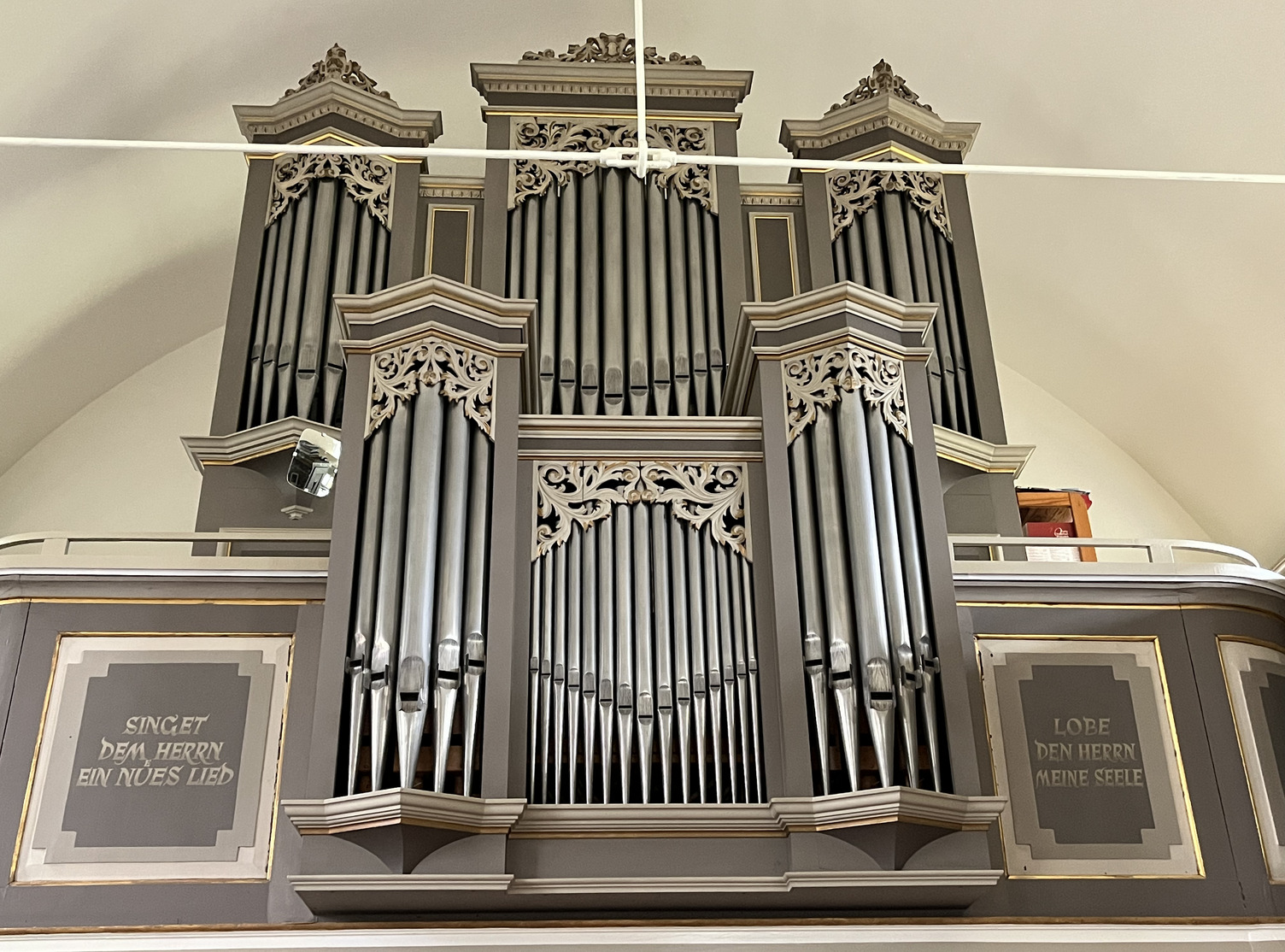 Ansicht der Orgel mt grauer Vertäfelung. Zwei Schriften sind neben der Orgel angebracht: "Singet dem Herrn ein nües Lied" und "Lobe den Herrn meine Seele"