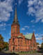 Eine Backsteinkirche mit hohem, spitzen Turm steht im Vordergrund, umgeben von herbstlichen Bäumen. Der Himmel ist blau mit einigen Wolken. Die Kirche hat große Fenster und einen Kupferdach. Der Eingang ist im gotischen Stil gestaltet.